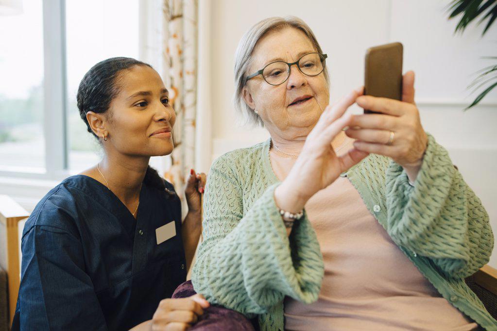 Smiling female caretaker looking at senior woman using mobile phone in nursing home