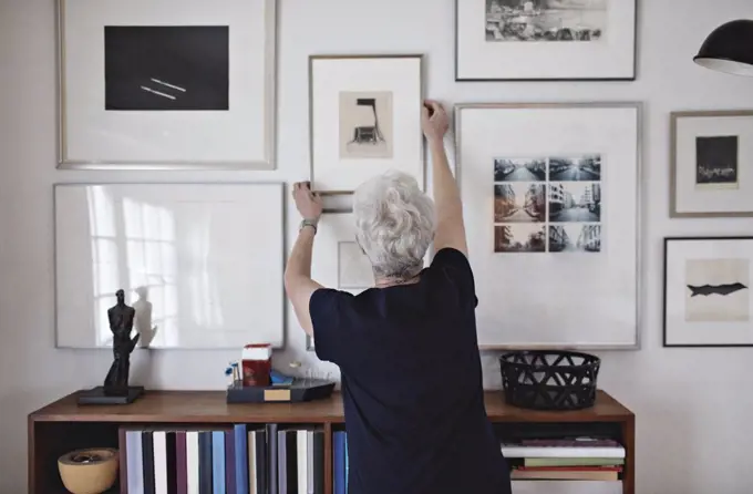 Rear view of retired senior woman adjusting picture frame on wall over bookshelf at home
