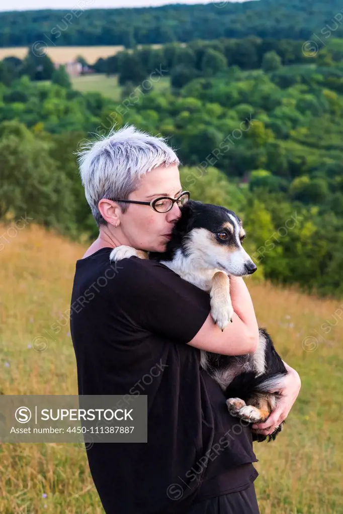 Woman with short grey hair wearing glasses standing on a hillside, holding dog.