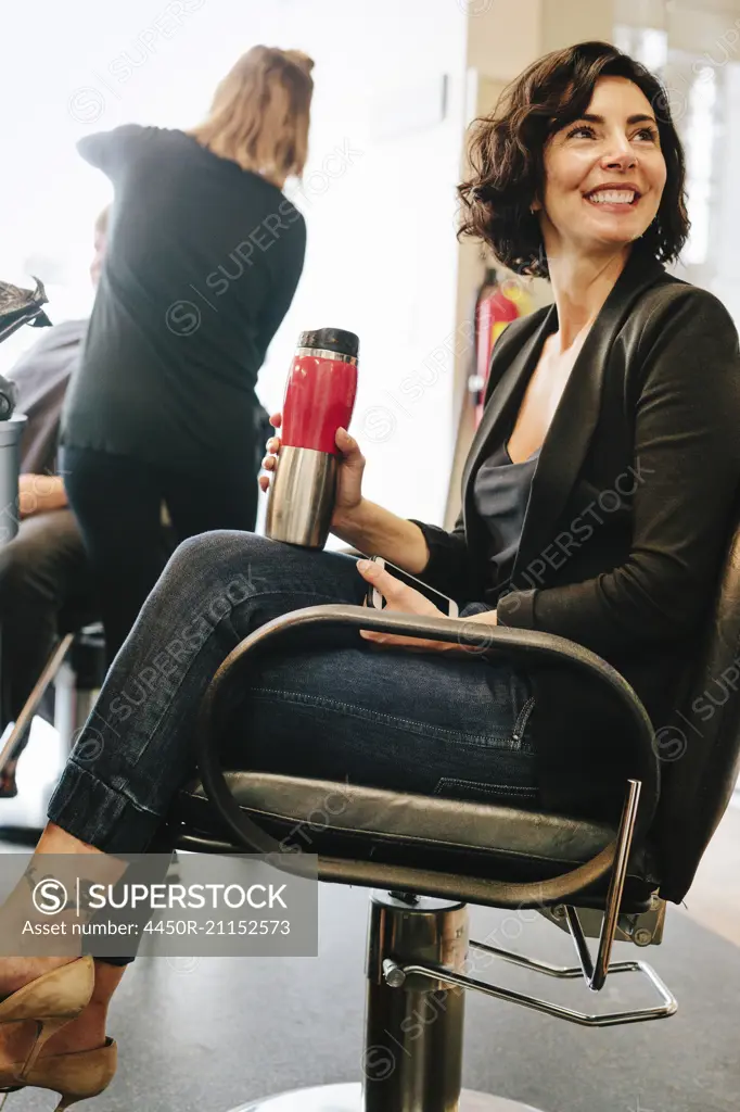 A woman client sitting in a hair salon chair, holding a mug of coffee.