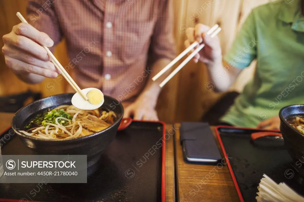 Two people, a Japanese man showing a Western man how to use chopsticks in a noodle shop.