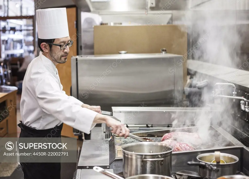 An asian chef working with fresh meat on the grill in a commercial kitchen,