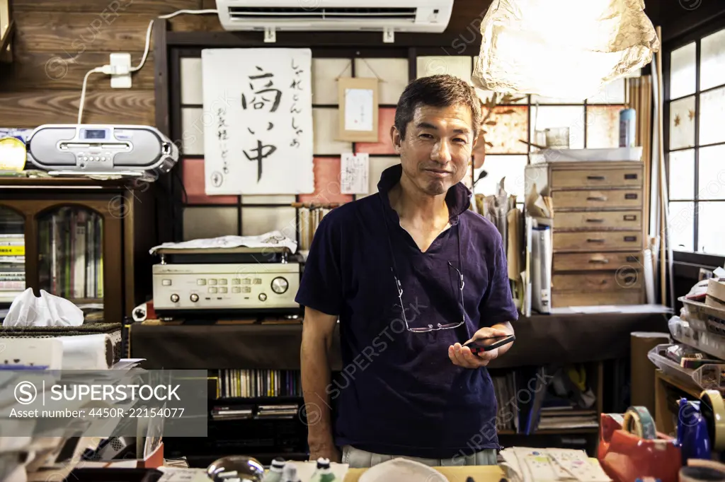 Japanese man standing in a Washi producing workshop holding a smart phone, smiling at camera.
