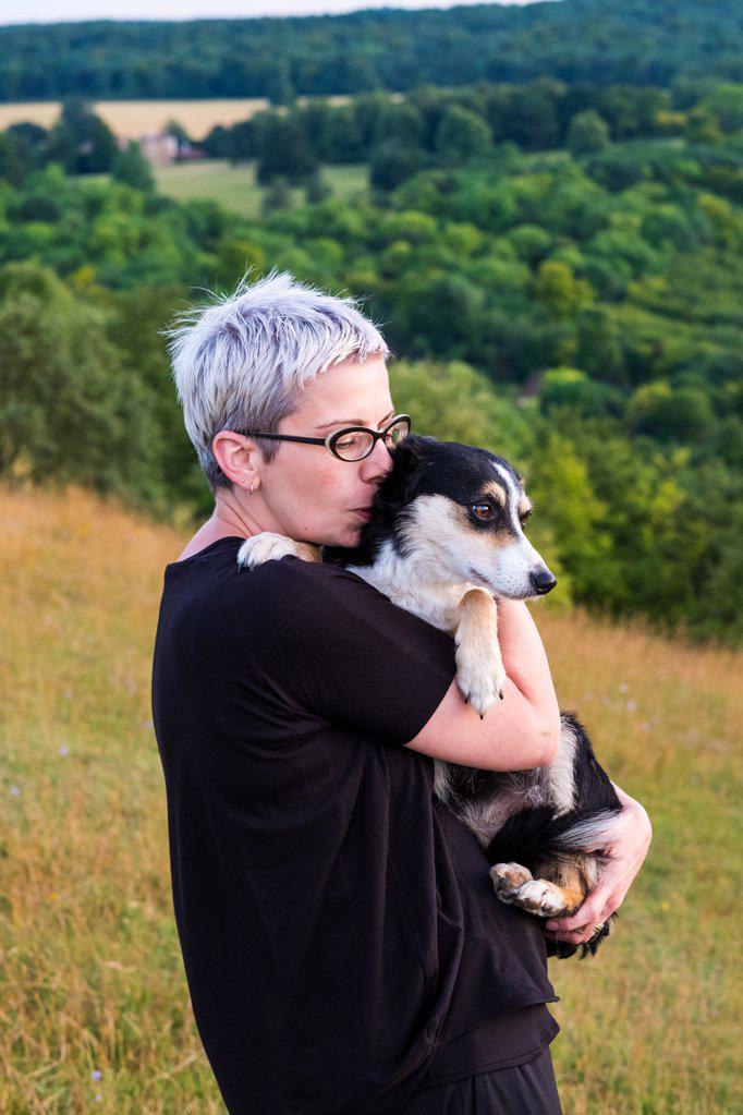 Woman with short grey hair wearing glasses standing on a hillside, holding dog.