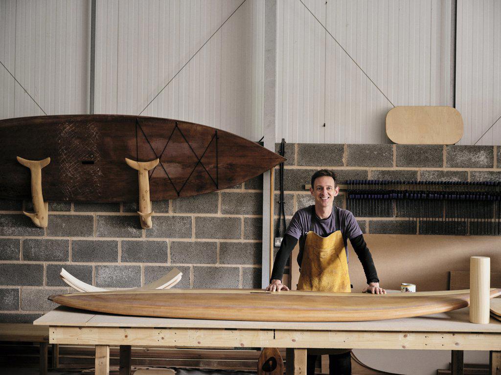 Portrait of man standing in front of hand made wooden paddleboard in workshop