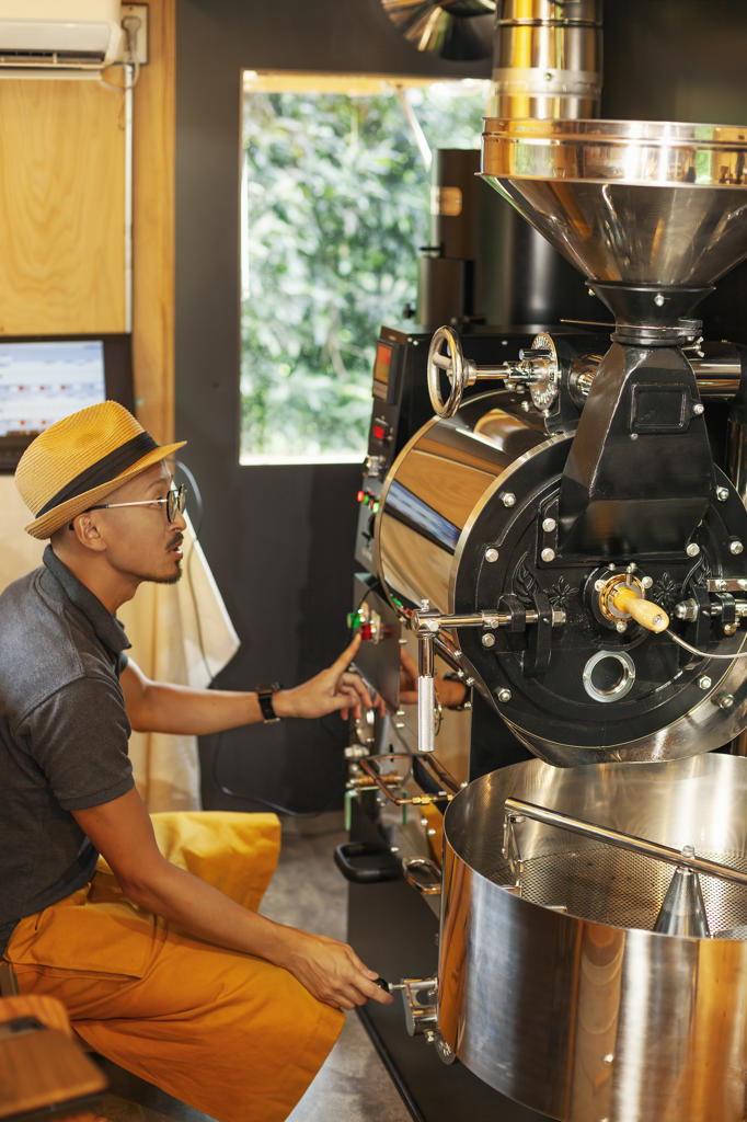 Japanese man wearing hat and glasses sitting in an Eco Cafe, operating coffee roaster machine.