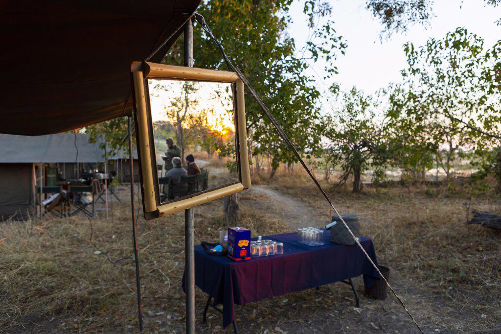 A safari camp, tents and drinks table, a large mirror on a tent pole, three people at sunset 