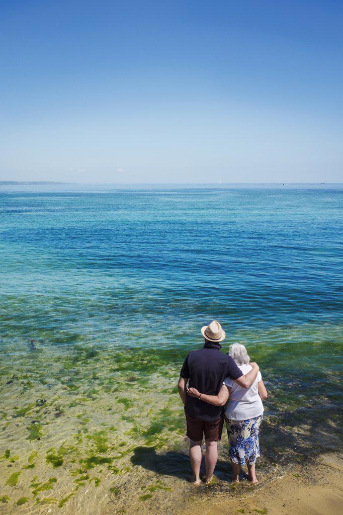 Rear view of elderly couple standing with arms around shoulders looking out to sea across the vivid clear blue water.