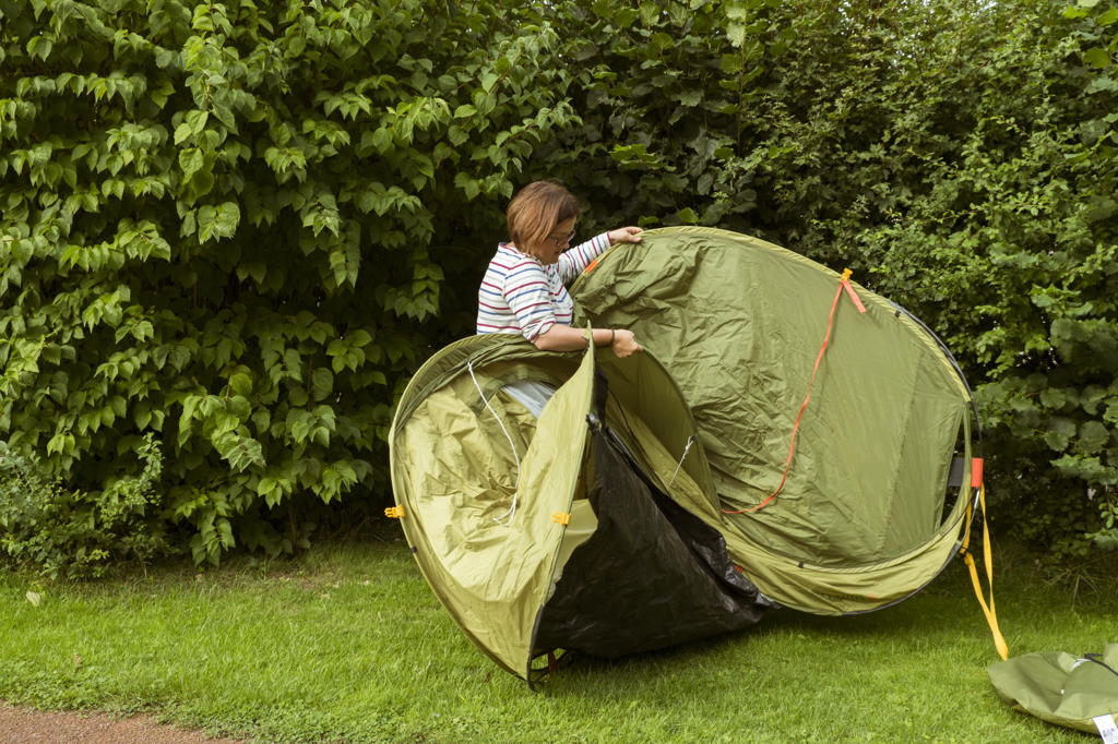 A woman putting up a green pop up tent in a sheltered spot by a tall hedge.