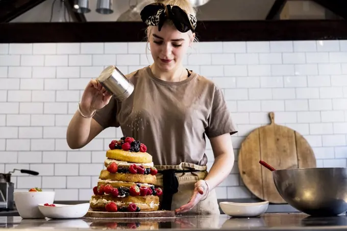 A cook working in a commercial kitchen sprinkling icing sugar over a layered cake with fresh fruit.