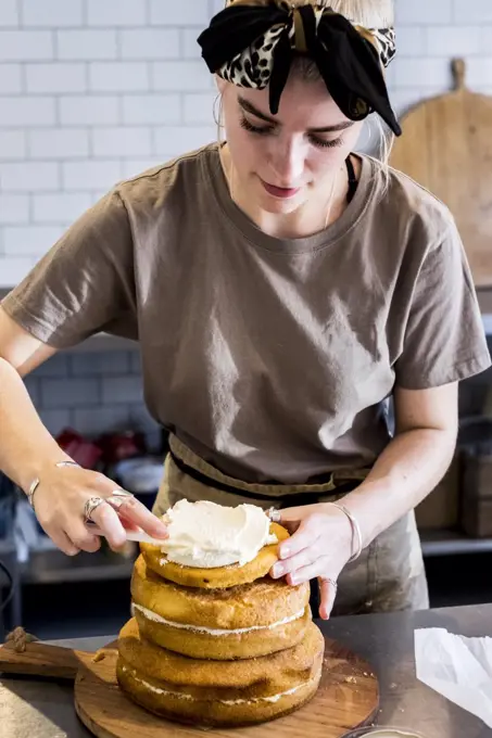 A cook working in a commercial kitchen assembling a layered sponge cake with fresh cream.