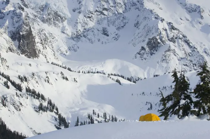 A small yellow tent pitched in deep snow on a slope, view of the steep slopes of the mountains. 