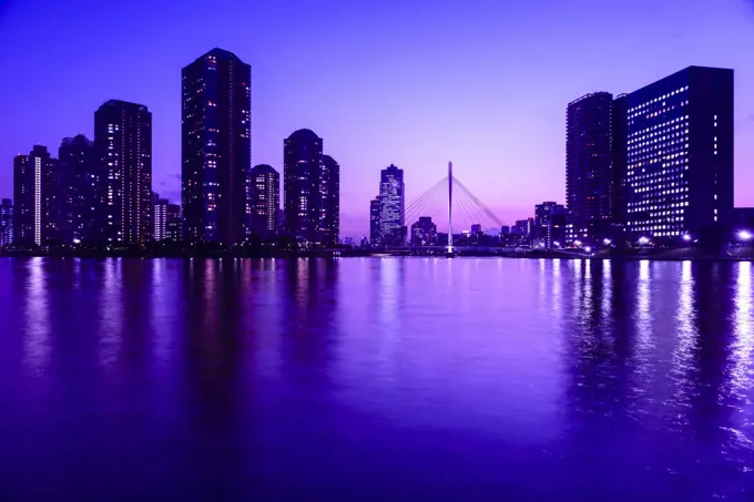 The city of Tokyo at night, the Sumida River, buildings silhouetted against the purple sky, reflections in the water.