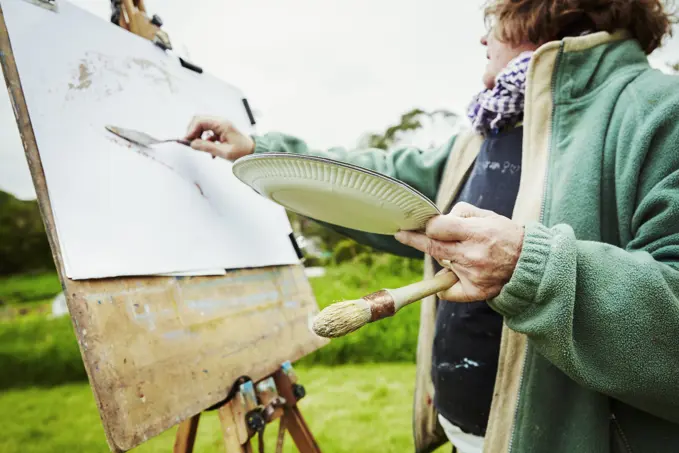 A woman artist working at her easel outdoors, applying paint with a handtool.
