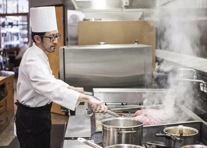 An asian chef working with fresh meat on the grill in a commercial kitchen,