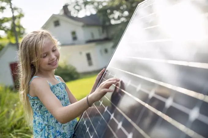 A young girl beside a large solar panel in a farmhouse garden.