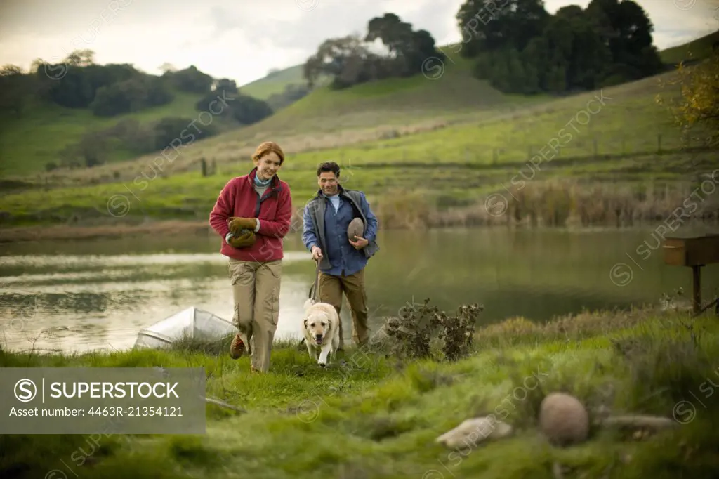 Mid-adult couple with their dog are carrying rocks beside a pond.