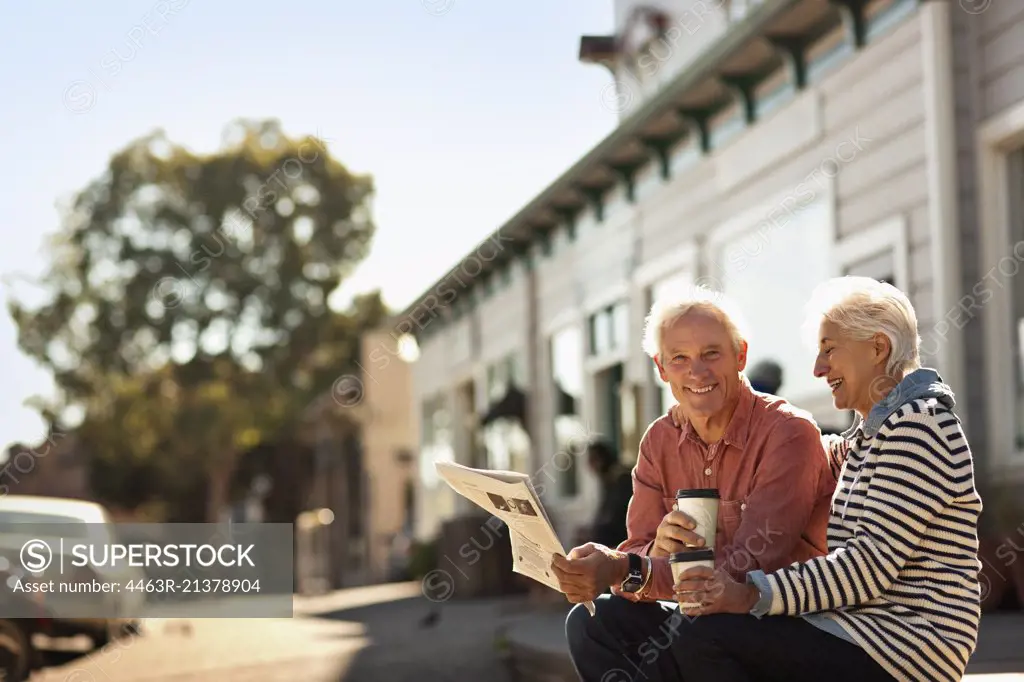 Smiling senior couple sit reading a newspaper and drinking coffee from takeaway cups. 