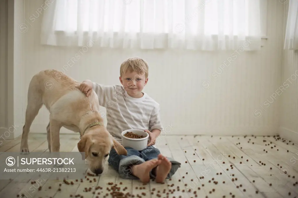 Young boy sitting on floor covered in dog biscuits with his dog. 