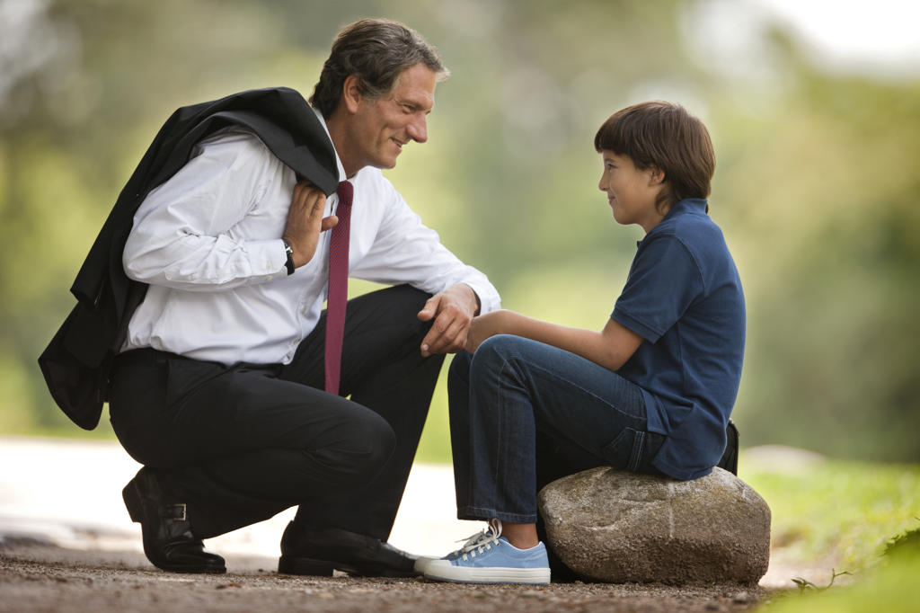 Businessman father crouches down and smiles at his young son who sits on a rock in a park. 