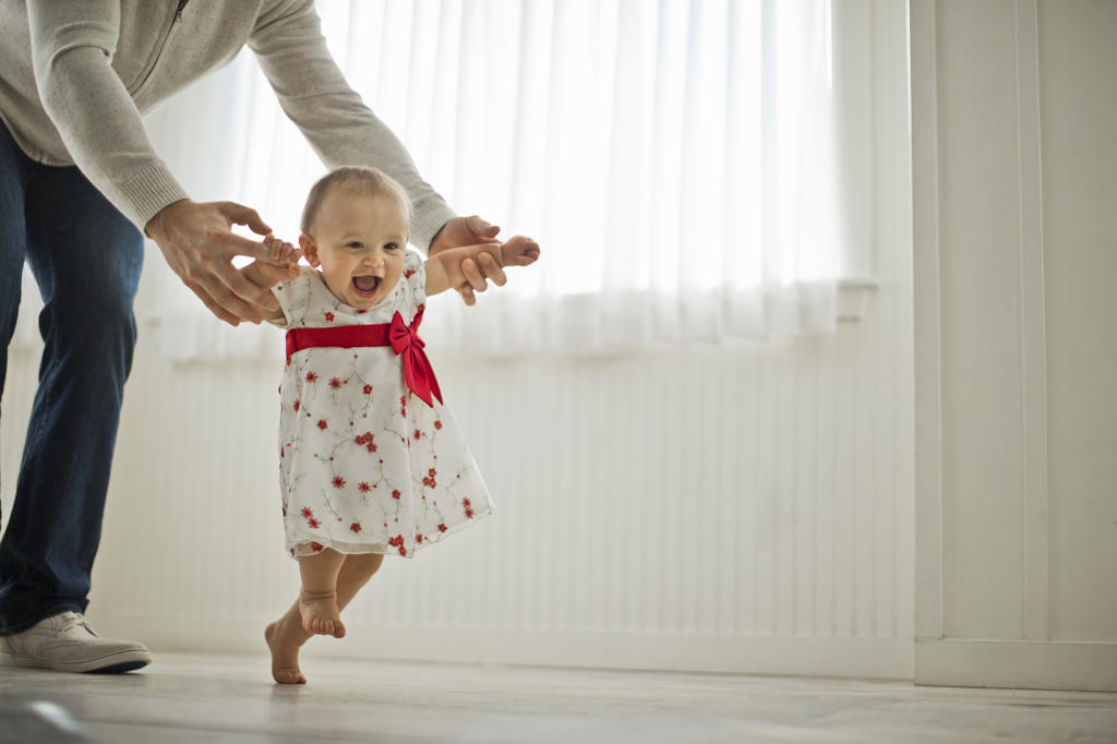 Sweet smiling baby girl in a floral dress takes her first steps assisted by her daddy.
