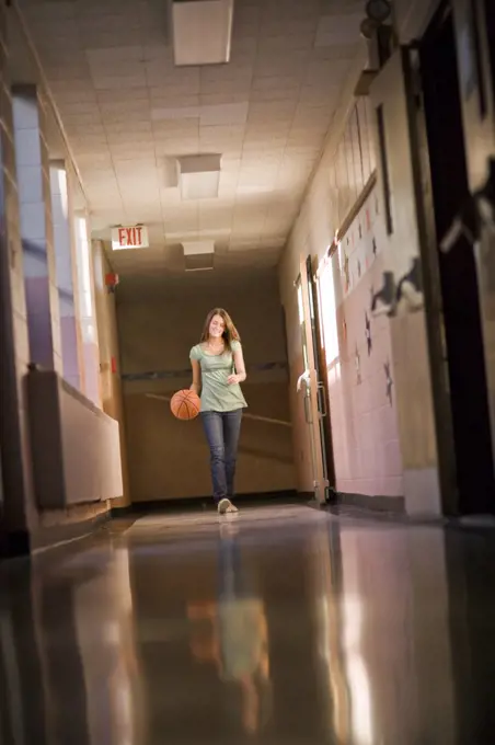 Teenage girl dribbling basketball in school corridor