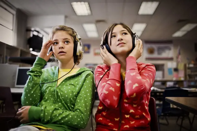 Two teenage girls sitting with headphones on in a school.
