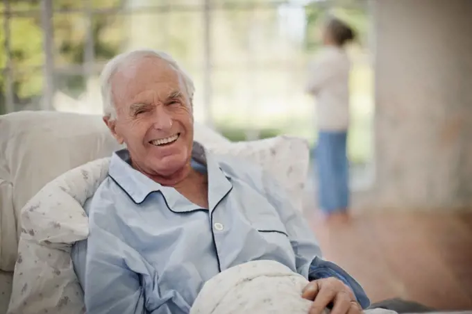 Portrait of a laughing senior man lying in bed with his wife standing in the background.