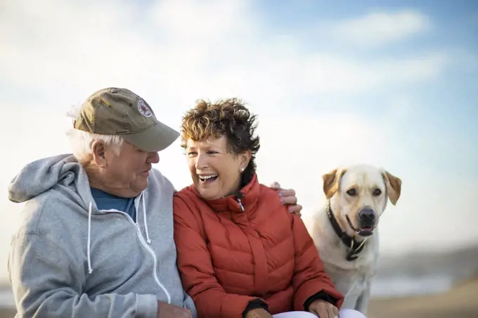 Happy senior couple sitting with their dog on a beach.