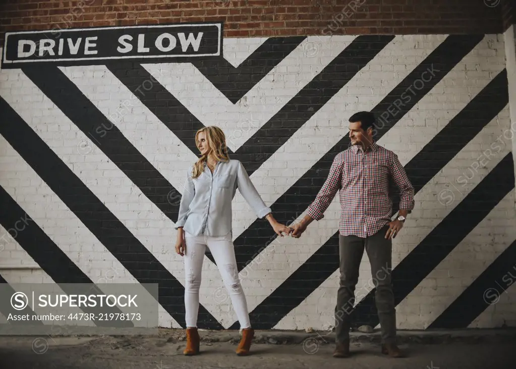 A man and woman standing holding hands in front of a black and white chevron board with the sign Drive Slow.