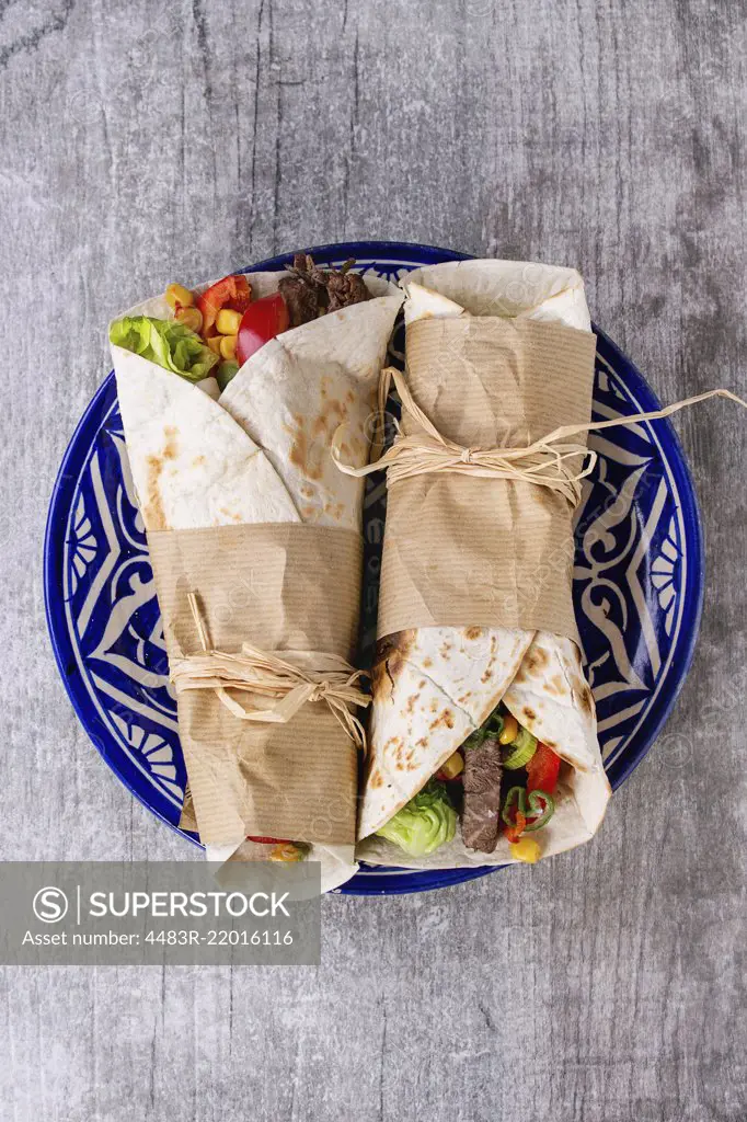 Mexican style dinner. Two papered tortillas burrito with beef and vegetables served on blue ornamental ceramic plate over white wooden background. Flat lay. With copy space