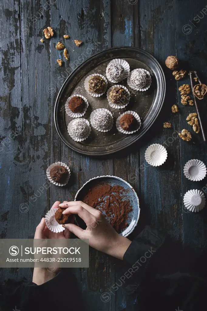 Child hands make homemade dark chocolate truffles with cocoa powder, coconut, walnuts, put on vintage tray over old dark blue wooden background. Top view, space. Rustic style