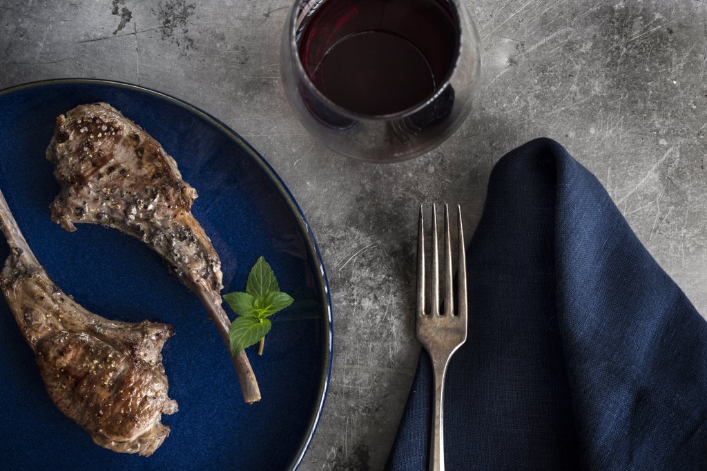 Roasted lollipop lamb chops on blue plate. Table setting with navy linen, red wine and rustic metal surface.