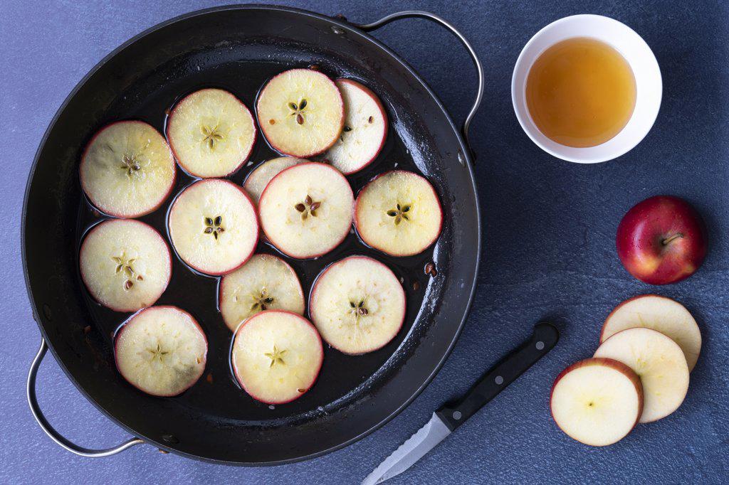 Sliced apples in a pan of honey syrup with a bowl of honey, a knife and a sliced apple.