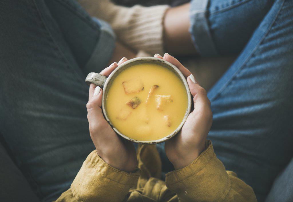 Flat-lay of female in yellow shirt and jeans sitting and keeping mug of Fall warming yellow pumpkin cream soup with croutons, top view. Autumn vegetarian, vegan, healthy comfort food eating concept