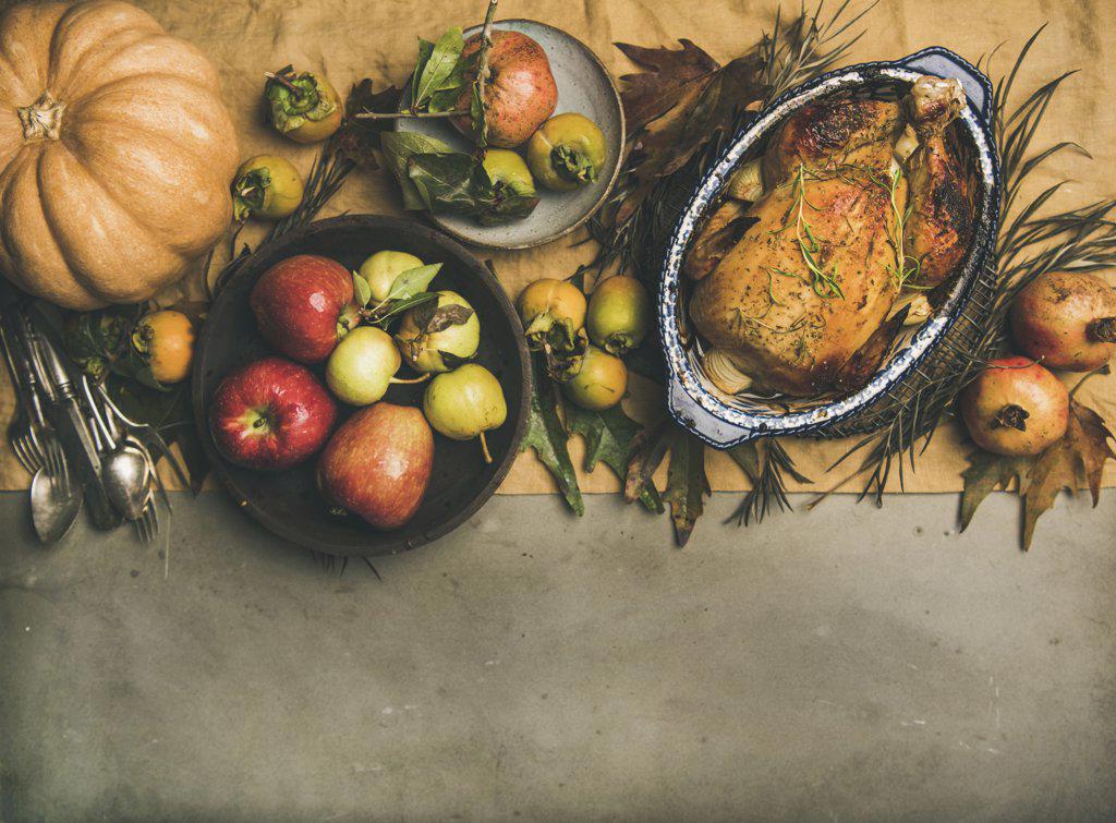 Thanksgiving dinner table. Flat-lay of roasted chicken or turkey, autumn fruit, pumpking, cutlery, leaves decoration over yellow linen table runner on grey concrete background, top view, copy space