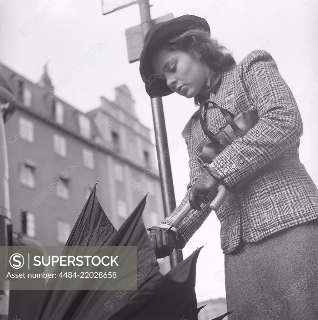 1940s woman in raincoat. Actress Viveca Lindfors, 1920-1995, is waiting at a bus stop and folds her umbrella together when it approaches. She is dressed in 1940s short chequered jacket and skirt, matching hat, handbag and gloves. August 1940. Photo Kristoffersson 159-20