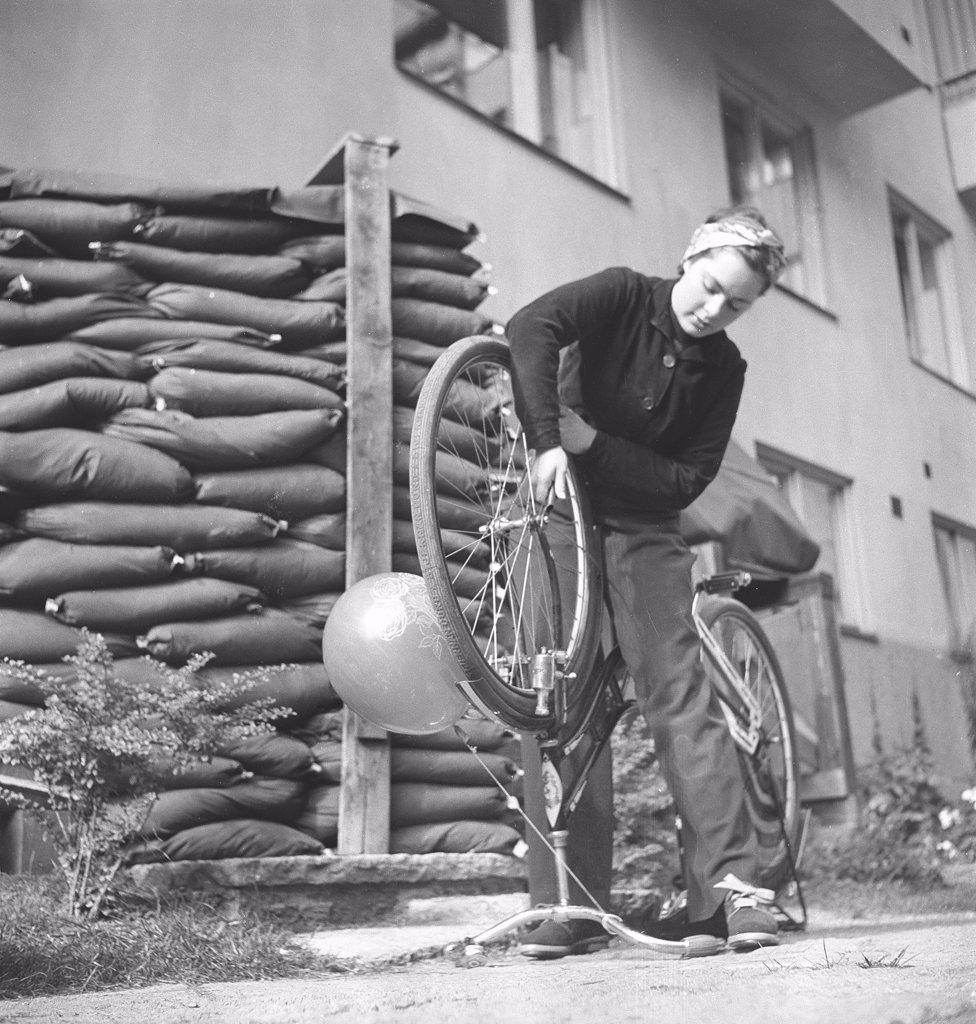1940s woman on a bicycle. Swedish actress Viveca Lindfors is repairing her bicycle. Notice the sand bags placed in front of the apartment building and the entrance to the basement shelter. Sweden August 1940. Photo Kristoffersson 159-32