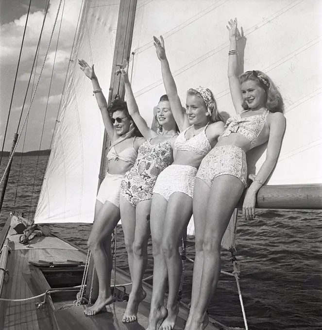 1940s sailing boat. Four young women are standing and leaning against the sails of the boat waiving as you should when you meet another boat. They are wearing the typical 1940s bathingsuit and bikini with high waist. Sweden 1946 Photo Kristoffersson ref AC100-4