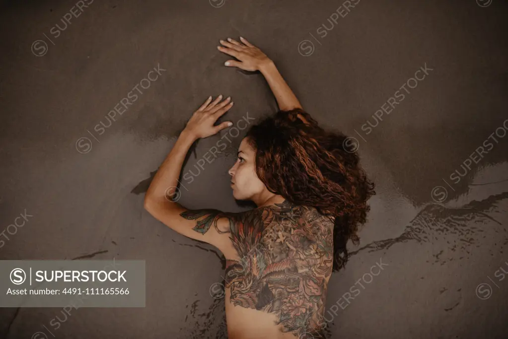 Naked female with tattooed back and shoulder lying on wet sand on seashore