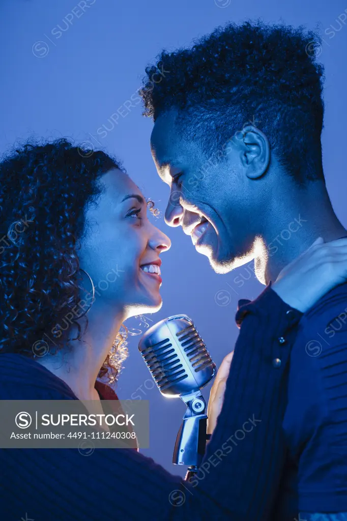 Side view of young African man and attractive woman holding vintage microphone and embracing in blue light