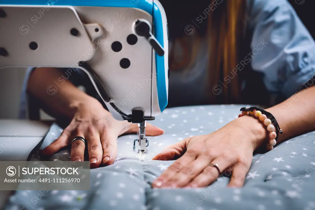 Crop view of hands of female tailor sewing on machine while sitting at table of workshop