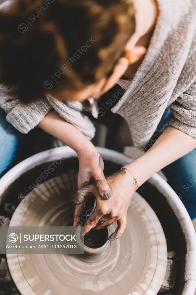 Anonymous female potter with dirty hands making clay pot on wheel in workshop