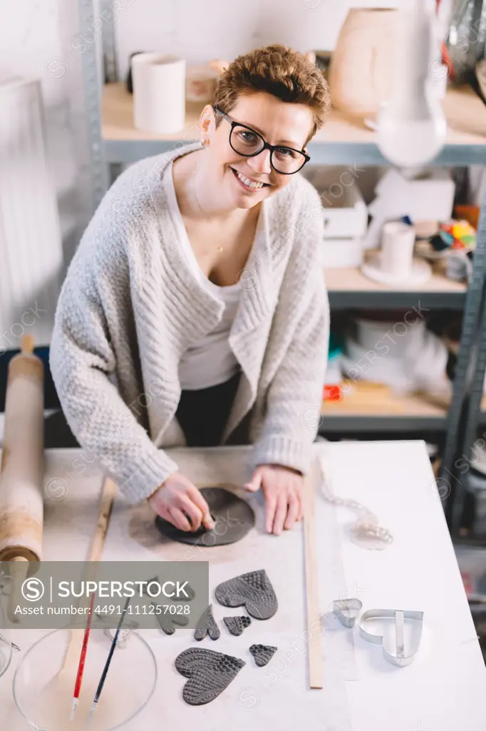 Adult woman in glasses cheerfully smiling and looking at camera while working with clay on table in potter workshop