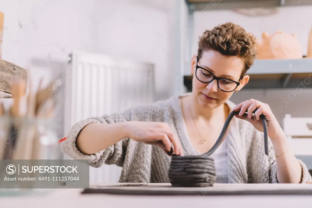 Adult female potter putting soft clay strip on top of vase while sitting at table in workshop