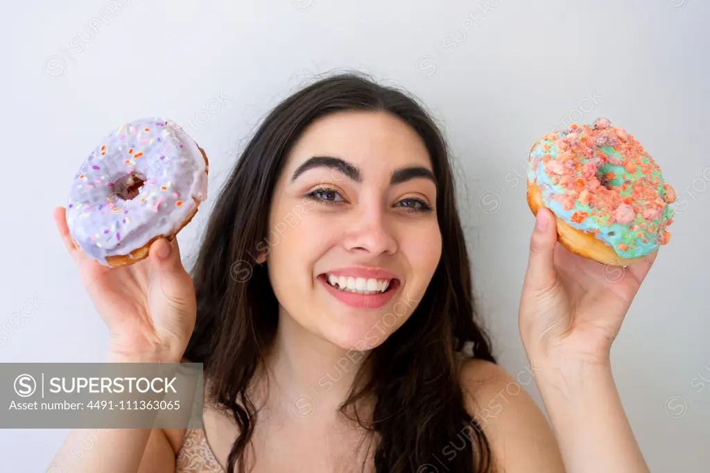 Carefree woman having fun and playing with glazed pastry with sprinkles while standing by white wall