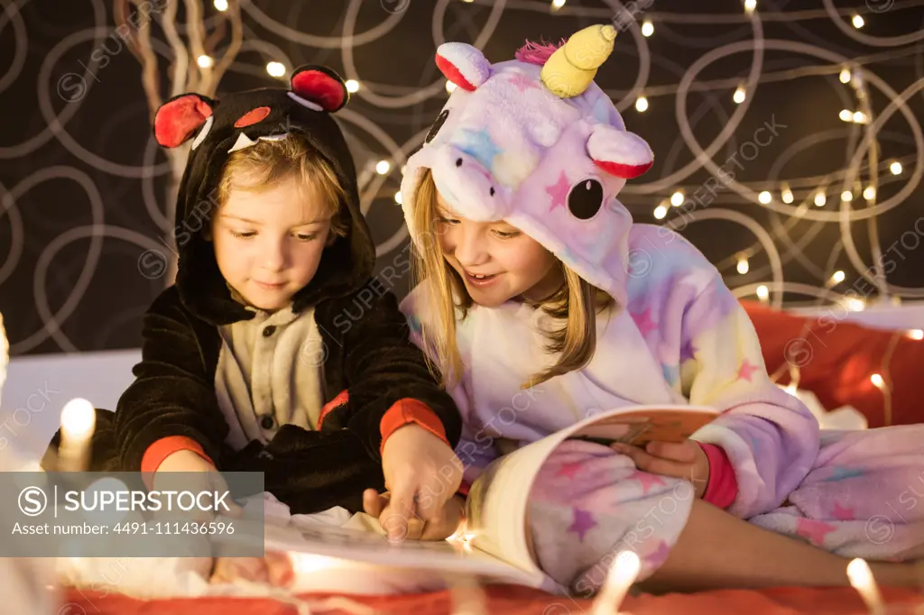 Cute little siblings in cozy kigurumi pajamas reading book with fairy tales while sitting together on bed decorated with Christmas lights