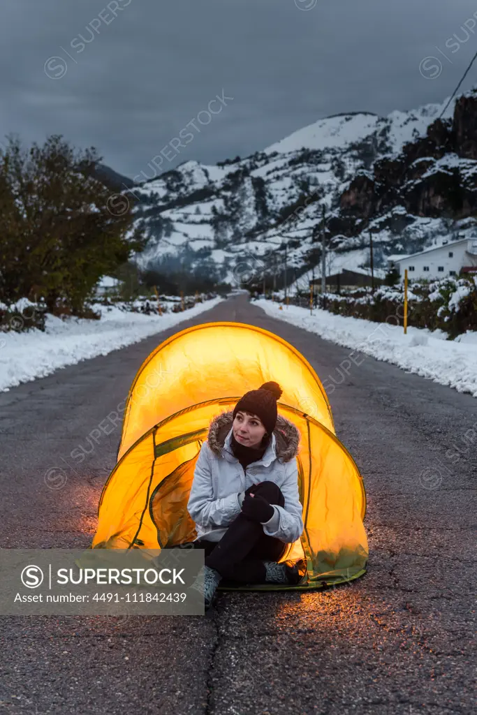 Woman in winter clothes sitting at entrance of tourist tent illuminated from inside standing in middle of asphalt road in mountain village at evening
