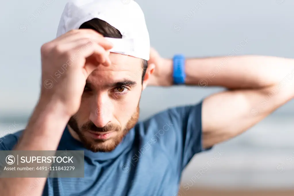 Sportsman in blue t shirt with hands behind head on white cap looking at camera with empty sandy seaside on blurred background