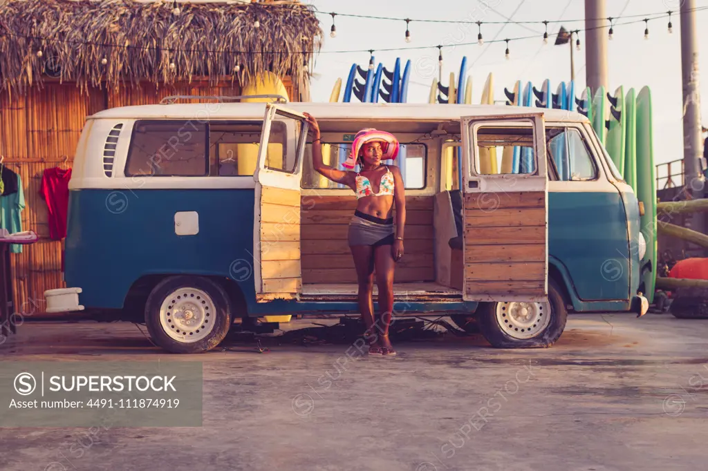Young African American female owner of local surf board rental wearing colorful bikini top and hat standing next to vintage van with row of surf boards in background;African American female standing next to retro van